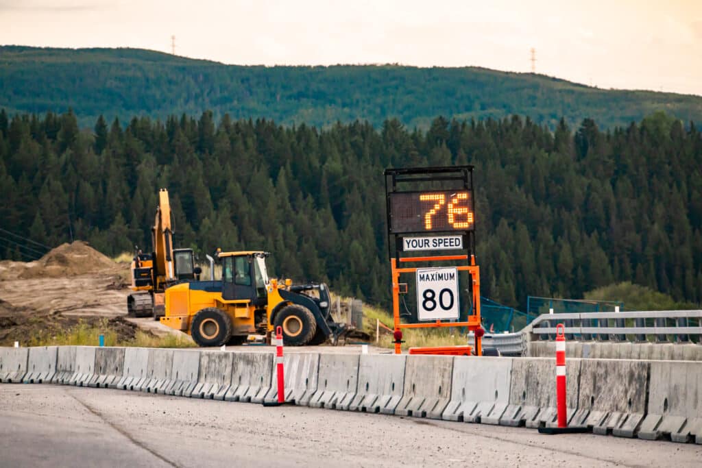 A construction zone set up on a highway with highway barricades, a traffic speed sign, and other construction safety products near the mountains. https://www.123rf.com/photo_135932197_radar-speed-sign-displays-vehicle-speed-on-variable-sign-your-speed-is-76-80km-maximum-limitation-ro.html?vti=m9zfg7nym1gqau1x8r-1-54