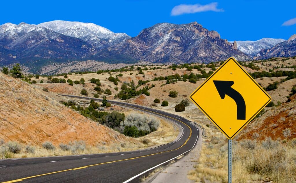 A yellow road sign meaning caution ahead as motorists take winding turns through a valley with a mountain in the background https://www.123rf.com/photo_20298945_curve-warning-sign-a-road-sign-alerts-motorists-to-a-curving-mountain-road-in-northern-new-mexico-.html 