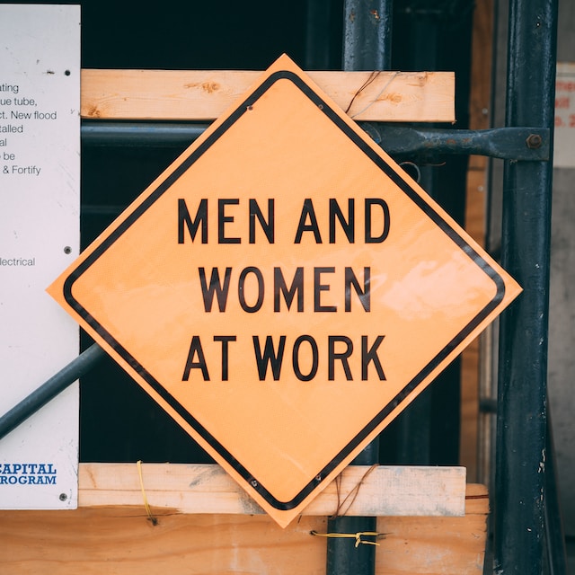 An orange type of construction sign that reads “Men and Women at Work”